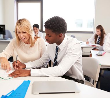 School Tutor Giving Uniformed Male Student One To One Tuition At Desk In Classroom